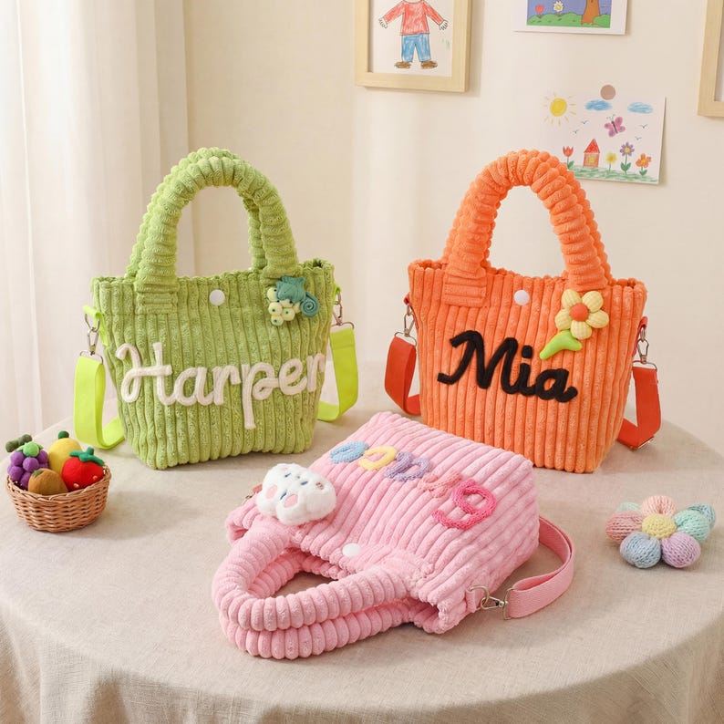 May include: Three corduroy tote bags in green, orange, and pink, each with a handle and a shoulder strap. The green bag says "Harper", the orange bag says "Mia", and the pink bag has a cloud and rainbow design.