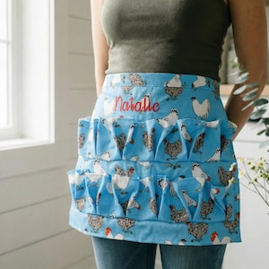 Pode incluir: Avental azul claro com um padrão repetido de galinhas em várias cores. O avental tem vários bolsos e o nome "Natalie" bordado em vermelho. O avental é projetado para coletar ovos.