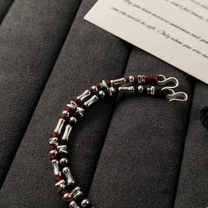 以下が含まれることがあります： シルバーカラーの金属ビーズとアクセントが付いた、2層のブレスレットのクローズアップ。 ブレスレットは赤と黒のコードが付いています。 手書きのテキストが書かれた白いカードが背景にあります。