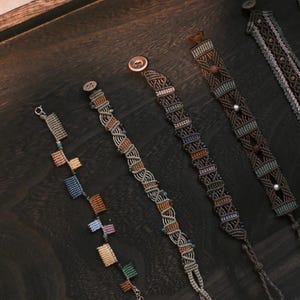 May include: A selection of macrame bracelets in diverse designs and colours, including brown, beige, blue, and orange. The bracelets are presented on a dark wooden surface, highlighting their intricate patterns and textures. Some have button fastenings.