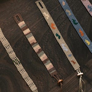 May include: Close-up of several woven friendship bracelets in various colors, including beige, brown, blue, and pink. Each bracelet features a unique pattern with decorative elements and a tassel or wooden bead closure. The bracelets are displayed on a dark wooden surface.