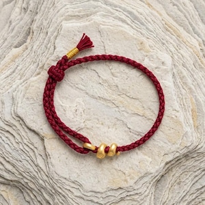 Könnte beinhalten: Ein Armband aus dunkelroter geflochtener Kordel mit zwei kleinen, matt goldfarbenen Metallringen, die auf die Kordel gefädelt sind. Das Armband verfügt über einen verstellbaren Schiebeknoten und ein kleines Quastenende mit goldfarbenem Garn.