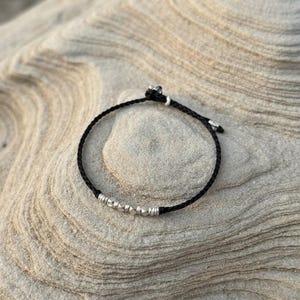Puede incluir: Una pulsera de cordón trenzado negro que presenta una serie de pequeñas cuentas metálicas de tono plateado dispuestas en línea. La pulsera incluye un mecanismo de cierre de nudo corredizo con dos pequeños acentos plateados.