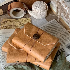 May include: Three brown, leather-effect journals with button and twine closures, stacked on a surface with vintage paper, lace trim, and grid tape. A wooden box is in the background, with other vintage items.