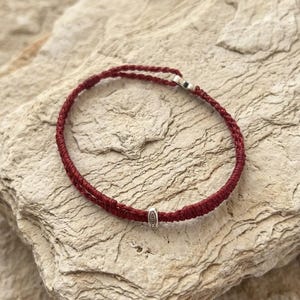 以下が含まれることがあります： 濃い赤色の編み込みコードブレスレット。魚のシンボルが刻印された小さな銀色の円筒形ビーズが付いています。このブレスレットは細い織りコードで作られており、ループの上部には金属製の留め具が見えます。