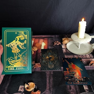 Op de afbeelding: Een groen tarotkaartendek met gouden folie accenten. De kaart die wordt getoond is De Dwaas, die een figuur in een narrenkostuum voorstelt die naar een klif loopt. De kaart ligt op een tafel met andere tarotkaarten, een kaars en decoratieve items.