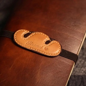 May include: A brown leather journal with a light brown leather bread-shaped closure. The word "Bread" is embossed on the closure, along with a small heart symbol. A black elastic band secures the journal.