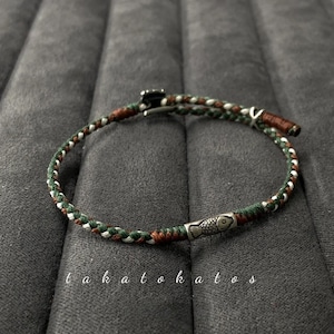May include: Braided bracelet with green, white, and brown threads, featuring a silver fish charm. The bracelet is secured with a metal clasp. The text "t a h a t o k a t o s" is written in cursive.