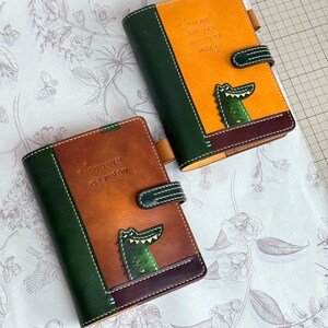 以下が含まれることがあります： クロコダイルのデザインの革製ジャーナル2冊。1冊は茶色で「Everyday is a good day」の文字入り。もう1冊はオレンジ色で「You are someone's reason to smile」の文字入り。どちらも緑色のアクセントが付いています。