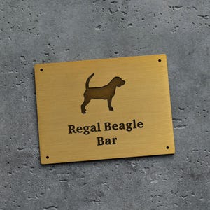 May include: A rectangular brass sign with the words "Regal Beagle Bar" and a silhouette of a dog. The sign has four small holes in each corner. The dog silhouette is brown.