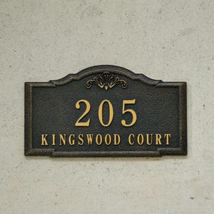 May include: A black and gold address plaque displaying the number 205 and the words KINGSWOOD COURT. The plaque features an ornate border and a floral motif at the top. The numerals and text are gold.