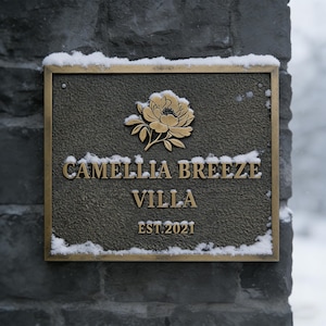 May include: A rectangular bronze sign with the words "CAMELLIA BREEZE VILLA" and "EST. 2021" in gold lettering. A gold floral design is at the top. The sign is mounted on a grey stone wall, with snow on top.