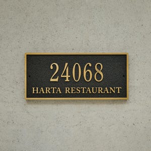 May include: A rectangular black sign with gold lettering and a gold border. The sign displays the number "24068" above the words "HARTA RESTAURANT". The sign is mounted on a light grey textured wall.