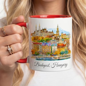 Op de afbeelding: Een witte keramische mok met een rode binnenkant en handvat, voorzien van een kleurrijke aquarelillustratie van de skyline van Boedapest, Hongarije, inclusief historische gebouwen en een rivier, met de tekst Budapest, Hungary eronder.