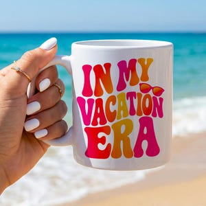 Könnte beinhalten: Ein weißer Keramikbecher mit dem Text IN MY VACATION ERA in einer fetten, wellenförmigen, mehrfarbigen Schrift in Pink, Orange und Gelb. Eine kleine Grafik einer roten Sonnenbrille befindet sich neben dem Wort VACATION auf dem Becher.