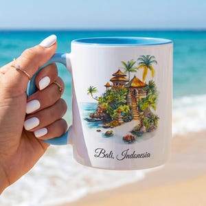 Pode incluir: Uma caneca de cerâmica branca com interior e alça azul-claro, apresentando uma ilustração colorida em aquarela de um templo num penhasco em Bali, Indonésia, com o texto Bali, Indonesia impresso em letras pretas abaixo da arte.