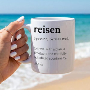Könnte beinhalten: Ein weißer Keramikbecher mit dem schwarz aufgedruckten Text 'reisen [rye-zuhn] German verb. to travel with a plan, a timetable and carefully scheduled spontaneity. Andreas'. Der Becher hat eine glatte Oberfläche und einen Henkel.