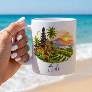 Könnte beinhalten: Ein weißer Keramikbecher mit einer farbenfrohen Illustration eines balinesischen Tempels, Palmen, Reisterrassen und einem Berg bei Sonnenuntergang. Das Wort Bali ist in schwarzer Schreibschrift unter dem runden Motiv aufgedruckt.