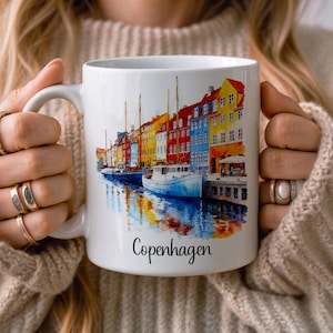 Op de afbeelding: Een witte keramische mok met een kleurrijke aquarelillustratie van de haven Nyhavn in Kopenhagen, Denemarken. Het ontwerp toont levendige rode, gele en blauwe gebouwen langs een kanaal met boten, met het woord Copenhagen eronder gedrukt.