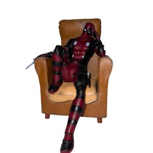 May include: A Deadpool action figure seated in a brown armchair. The figure is clad in a red and black suit and mask. A small silver sword is present. The chair has a worn look.