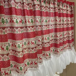 Puede incluir: Cortina navideña roja y blanca con un patrón festivo de renos, árboles y copos de nieve. La cortina tiene un borde inferior blanco con volantes, agregando un toque decorativo. La tela está estampada con rayas horizontales.
