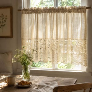 Op de afbeelding: Een beige linnen raamgordijn met een kanten rand, hangend voor een raam. Het gordijn is aan de bovenkant gerimpeld en heeft een decoratieve kanten rand aan de onderkant. Een houten tafel met een kopje, bord en bloemen staat ervoor.