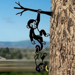 Op de afbeelding: Zwarte metalen apenbeeldhouwwerk dat aan een tak hangt. De sculptuur toont twee apen, waarvan één zich aan de tak vasthoudt en de ander een banaan vasthoudt. De achtergrond toont een boomstam en een vage landschap.