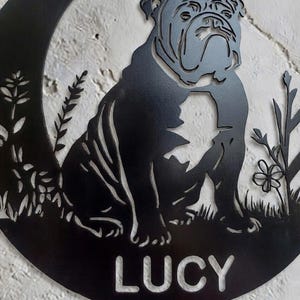 Puede incluir: Una pieza de arte mural circular de metal negro con una silueta recortada de un bulldog sentado entre elementos florales. El nombre LUCY est&aacute; recortado en letras may&uacute;sculas negritas en la parte inferior del marco circular decorativo.