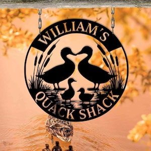 以下が含まれることがあります： 「WILLIAM'S QUACK SHACK」の文字が入った黒い金属製の丸い看板。アヒル、ガマ、水面のシルエットが描かれています。チェーンで吊り下げられています。