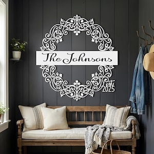 Puede incluir: Letrero con el nombre de la familia de metal blanco con un dise&ntilde;o de corona floral adornada. El letrero presenta el nombre de la familia "The Johnsons" y el a&ntilde;o "2025". El letrero est&aacute; montado en una pared gris oscuro sobre un banco de madera con cojines.