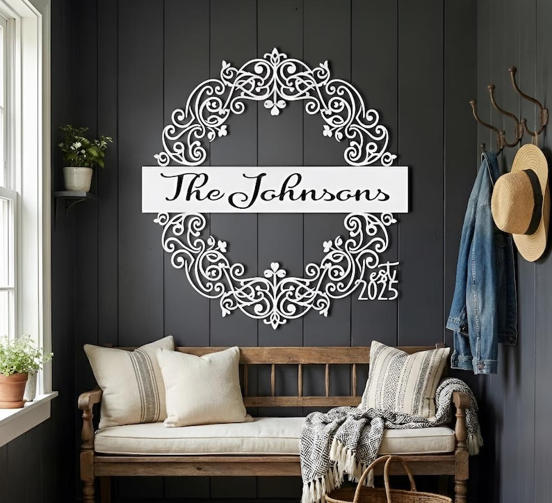 Puede incluir: Letrero con el nombre de la familia de metal blanco con un dise&ntilde;o de corona floral adornada. El letrero presenta el nombre de la familia "The Johnsons" y el a&ntilde;o "2025". El letrero est&aacute; montado en una pared gris oscuro sobre un banco de madera con cojines.