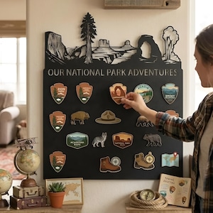 以下が含まれることがあります： OUR NATIONAL PARK ADVENTURESというタイトルの黒い金属製ウォールディスプレイ。上部には風景の切り抜きシルエットが施されています。ボードには、国立公園のアイコン、野生動物、ハイキング用品を描いた様々なマグネットやステッカーが配置されています。