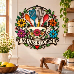 Peut inclure: Une enseigne murale circulaire représentant une tasse à thé colorée remplie d'une spatule, d'un fouet et de cuillères, entourée de fleurs et de feuilles. Le texte NANA'S KITCHEN est inscrit sur une bannière sous la tasse, avec un petit cœur rouge.