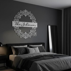 Puede incluir: Un letrero de nombre familiar de metal blanco con el texto "The Johnsons" y el a&ntilde;o "2025" dentro de un dise&ntilde;o de corona decorativa. El letrero est&aacute; montado en una pared gris oscuro sobre una cama con ropa de cama gris.