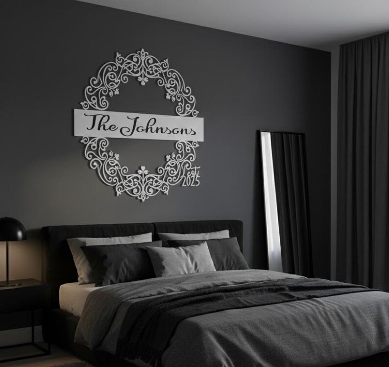 Puede incluir: Un letrero de nombre familiar de metal blanco con el texto "The Johnsons" y el a&ntilde;o "2025" dentro de un dise&ntilde;o de corona decorativa. El letrero est&aacute; montado en una pared gris oscuro sobre una cama con ropa de cama gris.