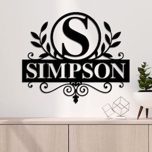 Puede incluir: Decoración de pared de metal negro con un círculo que contiene la letra "S" y la palabra "SIMPSON" debajo. El círculo está rodeado por dos ramas frondosas.