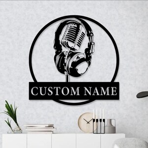 Op de afbeelding: Zwarte metalen wandkunst met een microfoon en koptelefoon in een cirkelvormig frame. Het ontwerp bevat de tekst "CUSTOM NAME" op een rechthoekige plaat eronder. Geschikt voor een muziekstudio of interieurdecoratie.