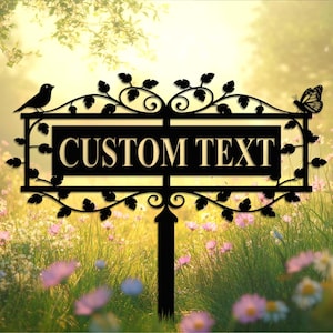 May include: Black metal garden sign with the words "CUSTOM TEXT". The sign has a decorative design with a bird on the left and a butterfly on the right. The sign is set in a garden with flowers.