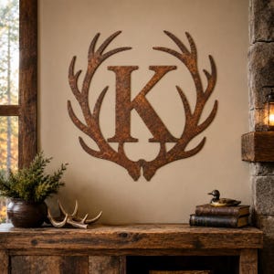 Op de afbeelding: Een aan de muur gemonteerd metalen monogrambord met de letter K in het midden van een cirkelvormig frame gevormd door twee symmetrische herten geweien. Het stuk heeft een getextureerde, verweerde, roestkleurige afwerking over het gehele oppervlak.