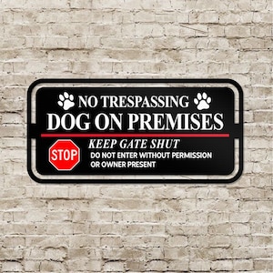 Op de afbeelding: Een zwart metalen bord met witte tekst die "No Trespassing Dog on Premises" luidt. Links op het bord staat een rood stopbord met witte tekst. Het bord luidt ook "Keep Gate Shut Do Not Enter Without Permission or Owner Present".