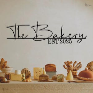 Może przedstawiać: Czarny metalowy znak z tekstem "The Bakery EST 2025" na białej ścianie. Na pierwszym planie stół z różnymi chlebami, bułkami i serami.