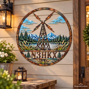 Puede incluir: Una pieza de arte mural circular de metal con un acabado rústico y desgastado. El diseño muestra un molino de viento central, una cordillera, pinos y un lago. El texto N3HKX aparece en la sección inferior, rodeado de tallos de trigo.