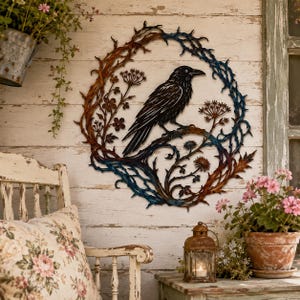 May include: A circular metal wall art piece featuring a black raven perched on a branch within a wreath of thorny vines and wildflowers. The metalwork displays a gradient of rust-orange and deep blue tones, creating a textured, layered appearance.