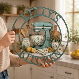 Op de afbeelding: Een cirkelvormig blauwgroen wandbord met de tekst Kelly's Kitchen, voorzien van een 3D-ontwerp van een keukenmixer, een mengkom met bloemmotieven, houten lepels, een garde, een snijplank, citroenen en kleine witte en gele bloemen.