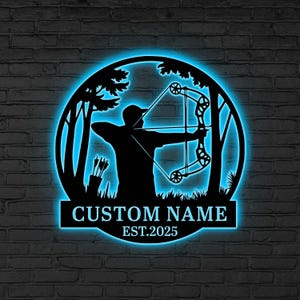 Op de afbeelding: Zwart metalen wandkunst met een boogschutter die een boog spant, geplaatst in een boslandschap, met een blauw omlijnde verlichting. De tekst "CUSTOM NAME EST.2025" staat onderaan het ontwerp.