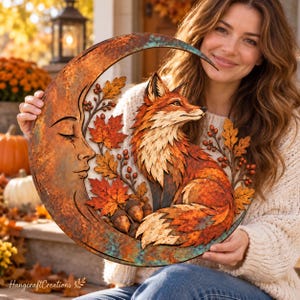 May include: A circular wooden wall art piece featuring a crescent moon with a carved face profile, a layered fox figure, autumn maple leaves, and acorns. The artwork displays a rustic, weathered finish with orange, brown, and teal patina tones.