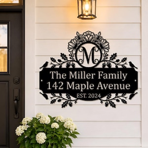 Op de afbeelding: Een zwart metalen wandbord met de letter M in een cirkelvormig frame, omringd door bloem-, blad- en eikelmotieven met vogel-silhouetten. De tekst luidt The Miller Family, 142 Maple Avenue en EST. 2024 in een strak wit lettertype.