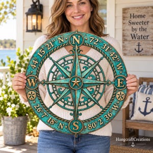 May include: A circular wall art piece featuring a compass rose design with a weathered teal and bronze finish. The outer ring displays cardinal directions N, S, E, W and coordinates 44 15 21 N, 72 31 42 W, 26 7 18 N, and 80 2 36 W in raised gold lettering.