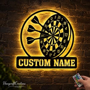 Op de afbeelding: Een rond wandbord van zwart metaal met een dartbordafbeelding met drie darts en een rechthoekig naamplaatje eronder met de tekst CUSTOM NAME. Het bord wordt verlicht door een warme gele LED-achtergrondverlichting en bevat een afstandsbediening.