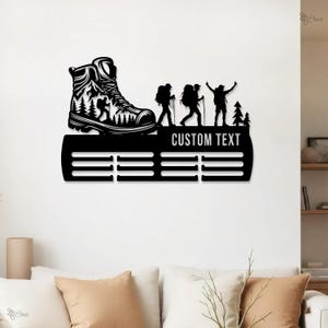 Puede incluir: Arte de pared de metal negro con la silueta de una bota de senderismo con un diseño de montaña y árboles. El arte incluye siluetas de excursionistas y el texto "CUSTOM TEXT" encima de barras horizontales para mostrar medallas. Una pieza decorativa.